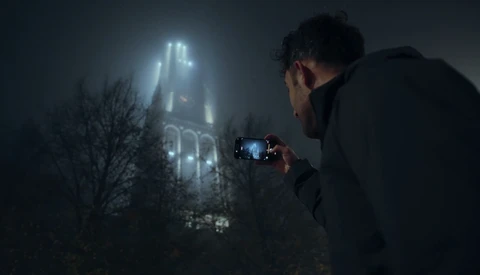 Photographer capturing a Gothic cathedral shrouded in fog at night with a smartphone.