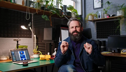 Bearded man in dark jacket sits at desk with laptop, gesturing while speaking in a plant-filled studio.