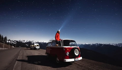 Person standing on roof of red and white vintage van at night, shining flashlight skyward with snow-capped mountains and starry sky in background.