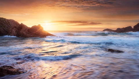 Waves crash on a rocky beach at sunset, with golden light illuminating the water and headlands.