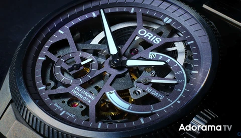 Close-up of an Oris luxury watch with exposed skeleton dial showing internal mechanisms and blue-toned case against dark background.