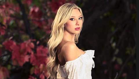 Woman with blonde wavy hair wearing an off-shoulder white ruffled top, photographed against a blurred background of pink flowering branches.