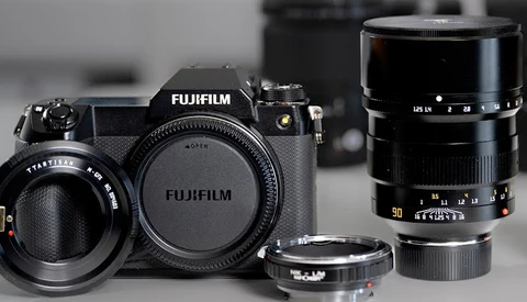 Fujifilm GFX medium format camera body with interchangeable lenses and adapter ring arranged on gray surface.