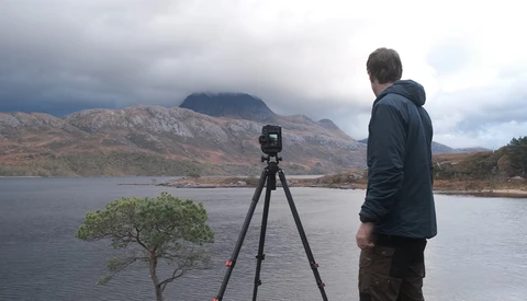 Photographer operating a tripod-mounted camera overlooking a mountain lake landscape.