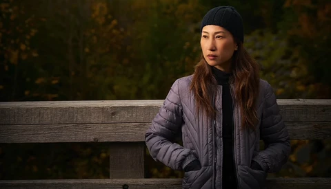 Woman in puffy jacket and black beanie sitting against wooden railing with blurred forest background.