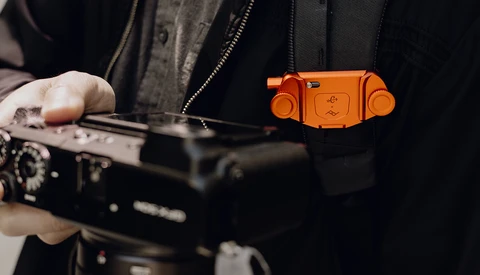 Photographer holding a camera with an orange camera strap accessory visible against dark clothing.