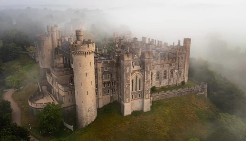 Medieval castle with multiple towers shrouded in mist, surrounded by forested hillside.