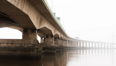 Long concrete bridge with supporting columns disappearing into thick fog over calm water.