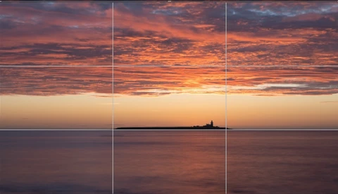 Seascape at sunset with a distant lighthouse silhouette centered on the horizon beneath dramatic orange and purple clouds.