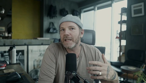 Man speaking into microphone in a home studio setup with casual attire and cap.