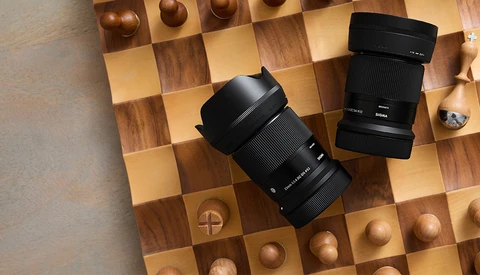 Two black camera lenses with lens hoods positioned on a wooden chess board.