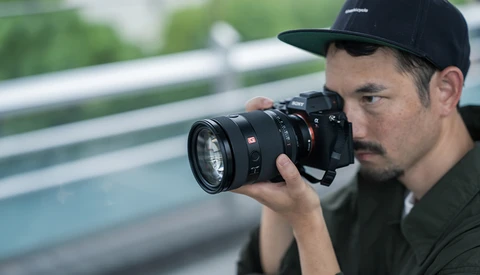 Photographer holding a mirrorless camera with a telephoto lens, wearing a black cap and olive shirt.
