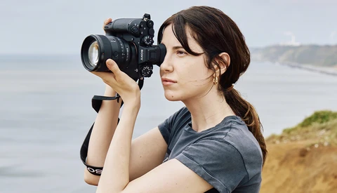Female photographer holding a DSLR camera with telephoto lens, positioned on a coastal cliff overlooking the ocean.