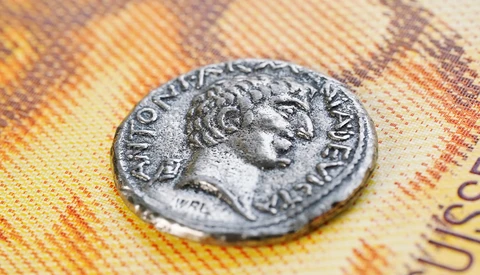 Ancient Roman coin with profile portrait resting on colorful euro banknotes.