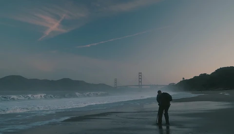 Person standing on wet beach at dusk with suspension bridge visible across the bay.