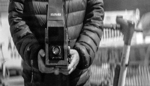 Hands holding a vintage rangefinder camera with distinctive striped quilted jacket sleeves visible.