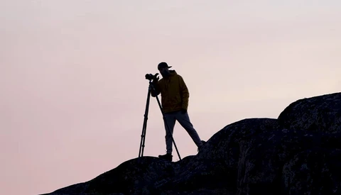 Silhouetted photographer standing on a rocky outcrop while looking through a tripod-mounted camera during twilight.