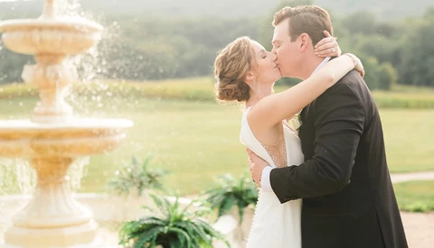 Bride and groom kissing by a fountain in a manicured garden setting.