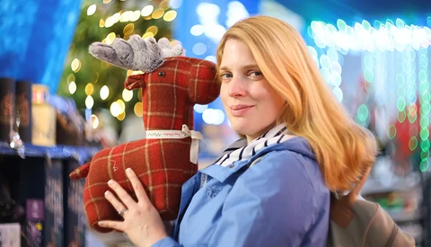 Woman holding red plaid stuffed dog toy indoors with holiday lights in background.