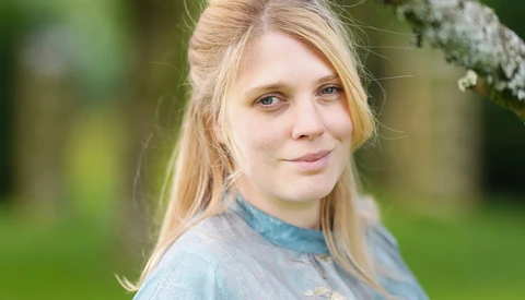 Young woman with blonde hair looking at camera in outdoor setting with green background.