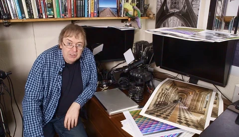 Photographer in a home studio workspace surrounded by monitors, camera equipment, and printed photographs.