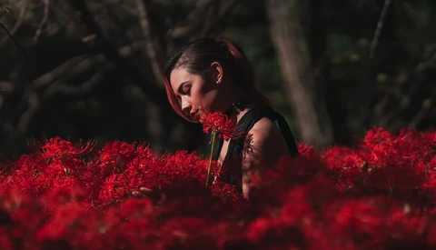 Woman with dark hair and closed eyes surrounded by vibrant red flowers in an outdoor setting.
