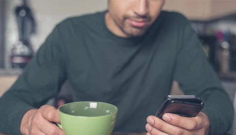 Man holding a green mug and smartphone, looking at phone screen while seated indoors.