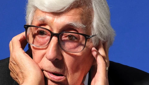 Older man with white hair and glasses against a blue background, hand near his face in a contemplative pose.