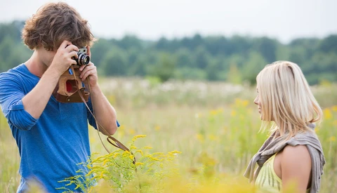 Young man with curly brown hair photographing a blonde woman in a sunlit field with vintage camera.