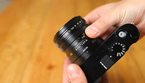 Hand holding a compact mirrorless camera with an attached prime lens against a warm wooden background.