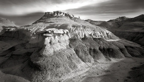 Dramatic black and white landscape of layered, eroded badlands with structures atop the highest ridge.
