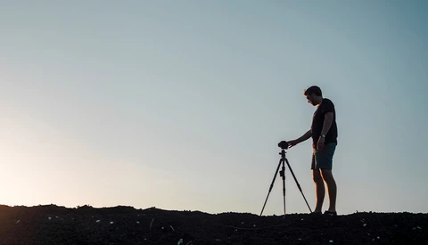 Silhouetted photographer operating a tripod-mounted camera against a clear sky at dusk.