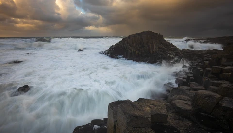 Waves crash over dark columnar basalt rocks jutting into turbulent sea at golden hour.