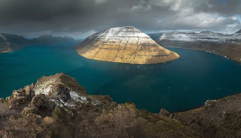 Snow-capped mountain with distinctive dome shape rising from turquoise fjord waters, viewed from rocky foreground.
