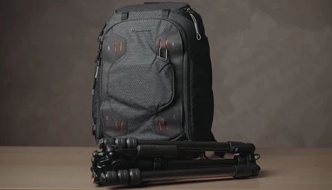 Gray camera backpack with red accent straps positioned next to a black tripod on a neutral surface.