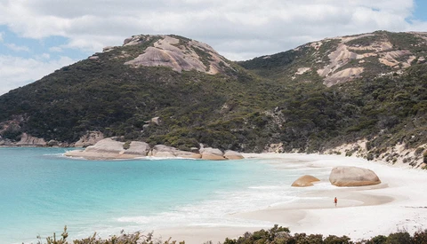Turquoise bay with white sand beach, large boulders, and forested hillside under partly cloudy sky.