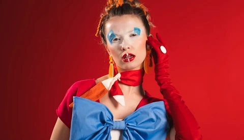 Portrait of a woman with blue eyeshadow and red lipstick against a red background, wearing a red jacket and blue bow.