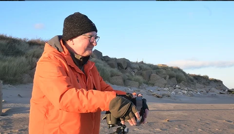 Photographer in orange jacket holding a camera on a beach with rocky cliffs in background.