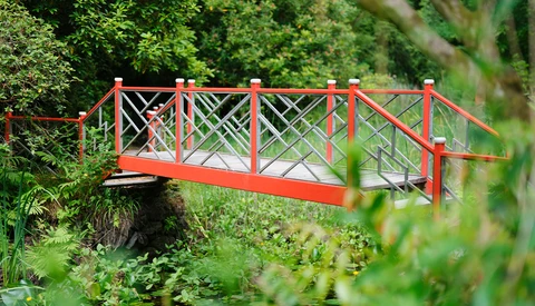 Bright red footbridge with X-pattern railings spanning a small stream in a lush garden.