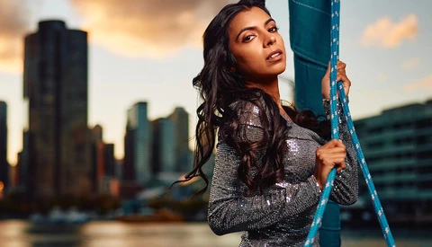 Woman in sequined top holding blue metal beam against urban skyline at sunset.