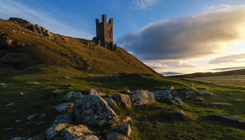 Medieval stone tower ruins on a hillside with golden hour sunlight and dramatic clouds.