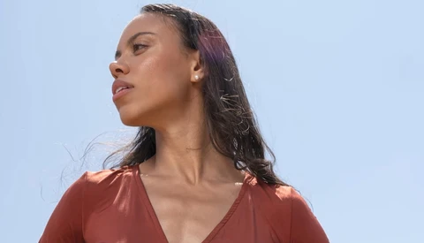 Woman with long dark hair wearing rust-colored top, photographed in profile against clear blue sky.