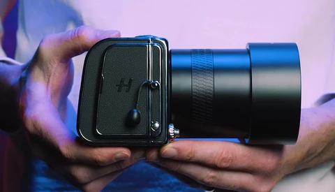 Hands holding a medium format camera with a telephoto lens against a purple-tinted background.