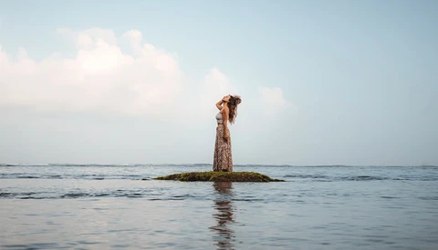 Woman in patterned dress standing on a wooden post jutting from calm shallow water under partly cloudy sky.