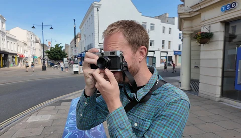 Man in checkered shirt holding vintage rangefinder camera to his face on a sunny urban street.
