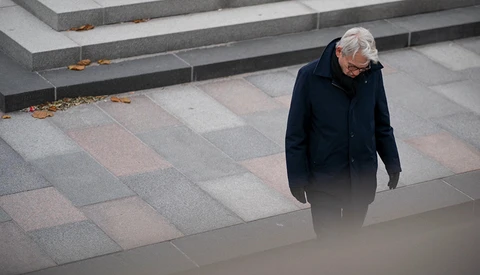Elderly person in dark coat walking alone across geometric paved plaza with stepped levels.