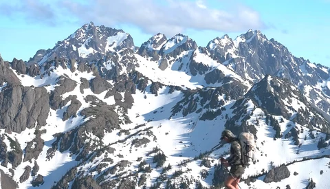 Snow-covered alpine peaks with forested slopes and exposed rock faces under partly cloudy skies.