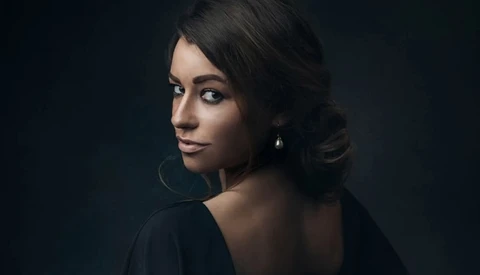 Woman with dark hair and hoop earrings looking over her shoulder against a dark background.