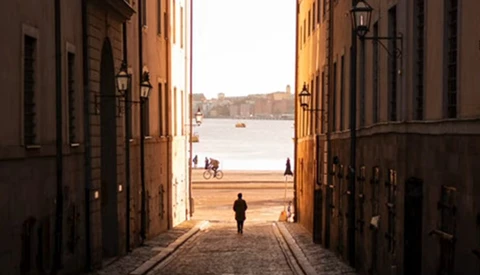 Narrow cobblestone alley between terracotta buildings framing a waterfront view with a solitary figure walking toward the water.