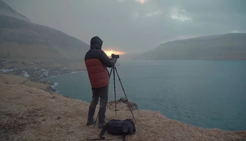 Photographer with tripod-mounted camera on hillside overlooking alpine lake at dusk.